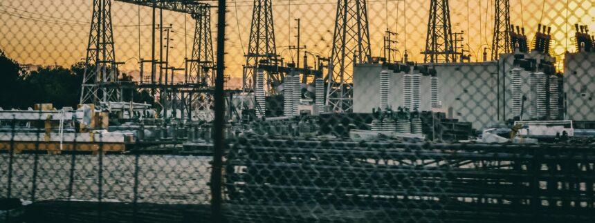 Industrial electric power substation photographed at sunset with fence in the foreground.